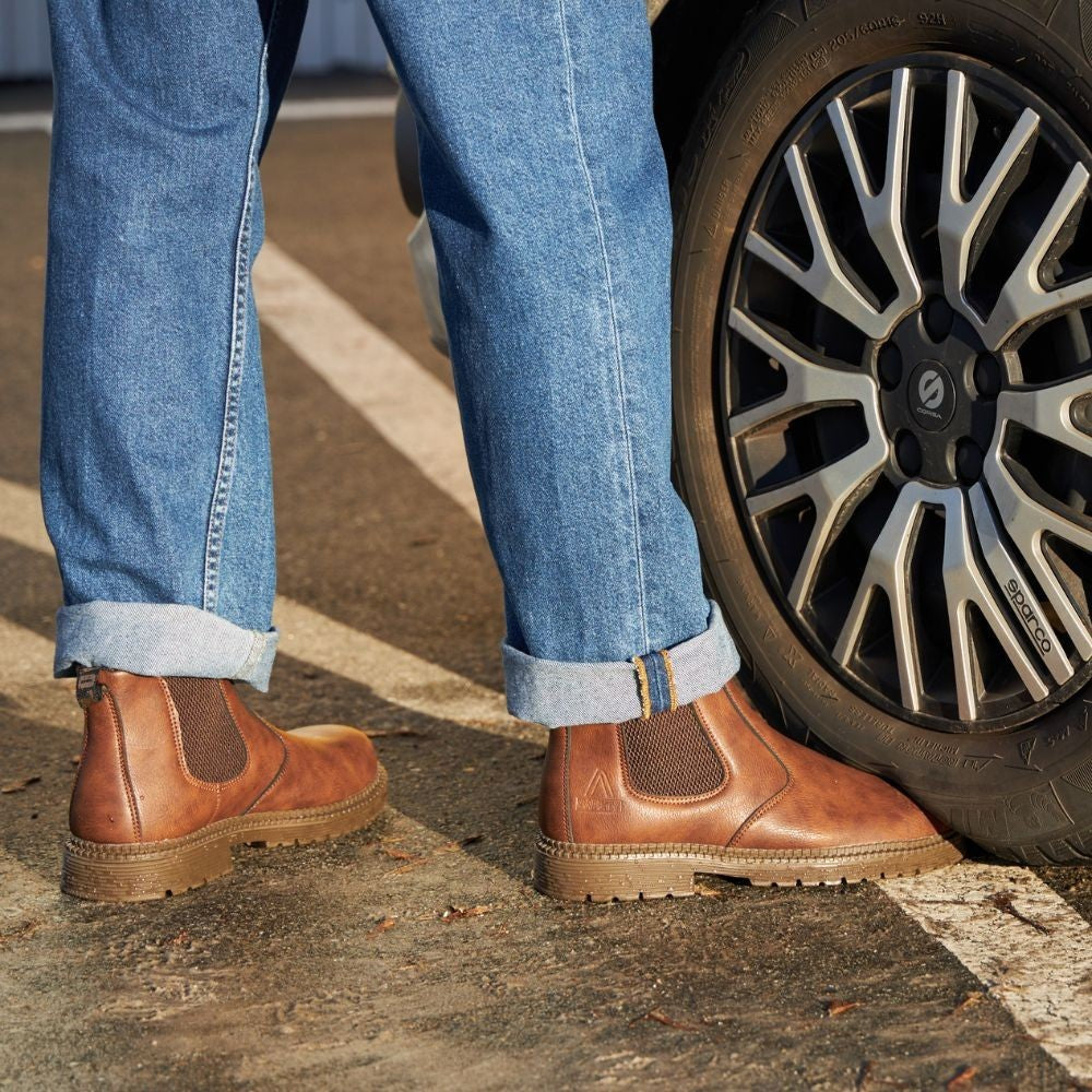 Braune Chelsea-Stiefeletten kombiniert mit hochgekrempelter Jeans neben Autoreifen