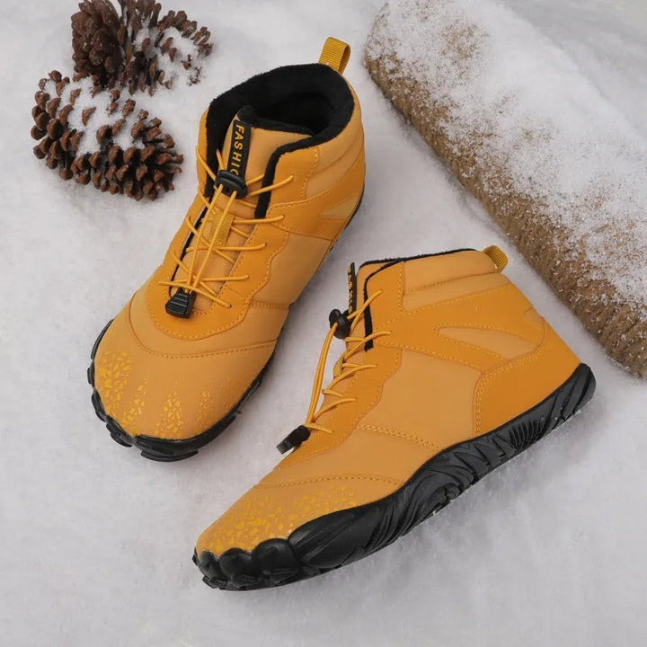 Senffarbene Barfußstiefel mit Profilsohle neben Tannenzapfen im Schnee platziert